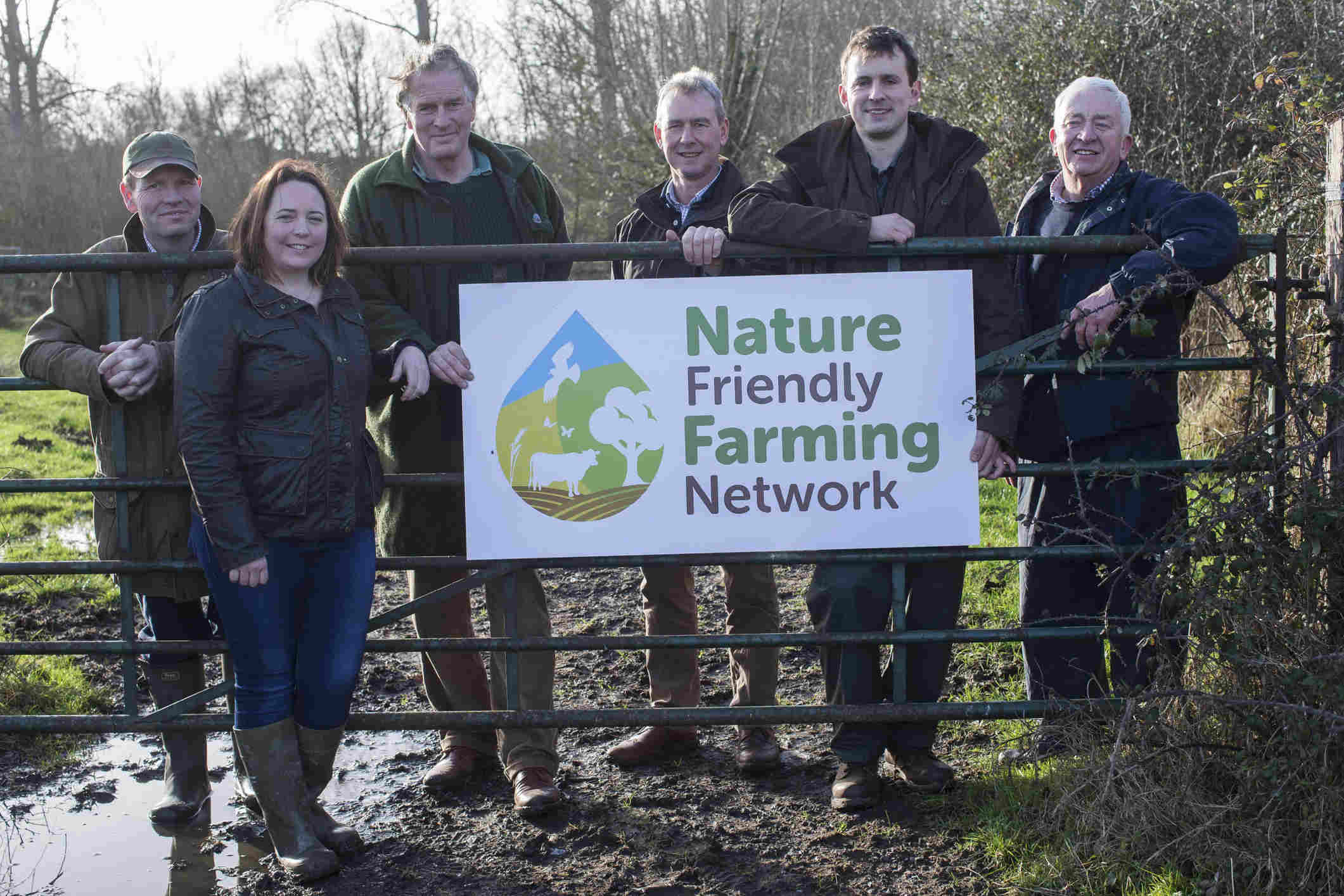 Five people leaning on a fence in a muddy field next to a sign: 'Nature Friendly Farming Network'.