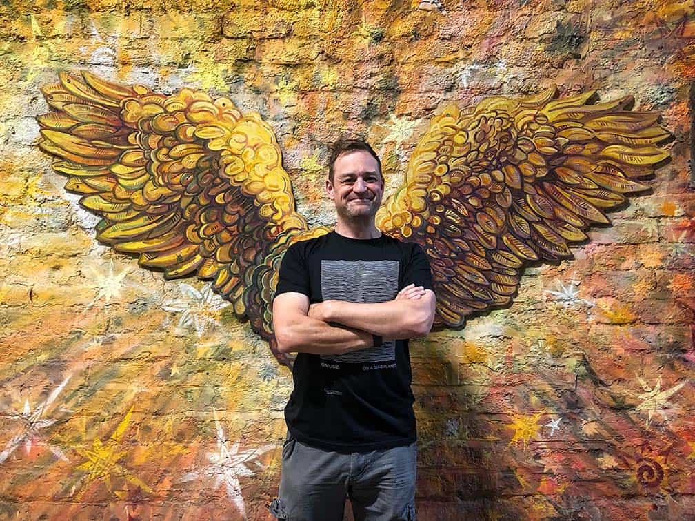 A white man is standing cross-armed wearing a black t-shirt and grey trousers. He is in front of a gold mural of a pair of angel wings.