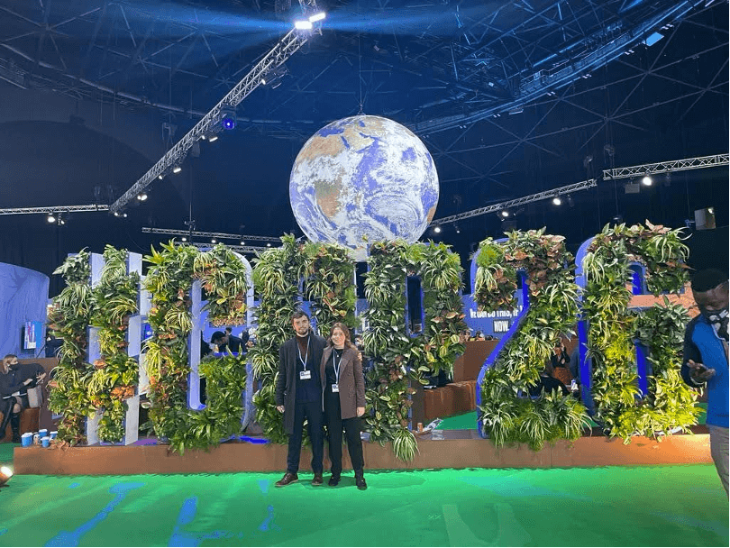Two people from Greenhouse Communications stand infront of giant COP 26 sign made of plants