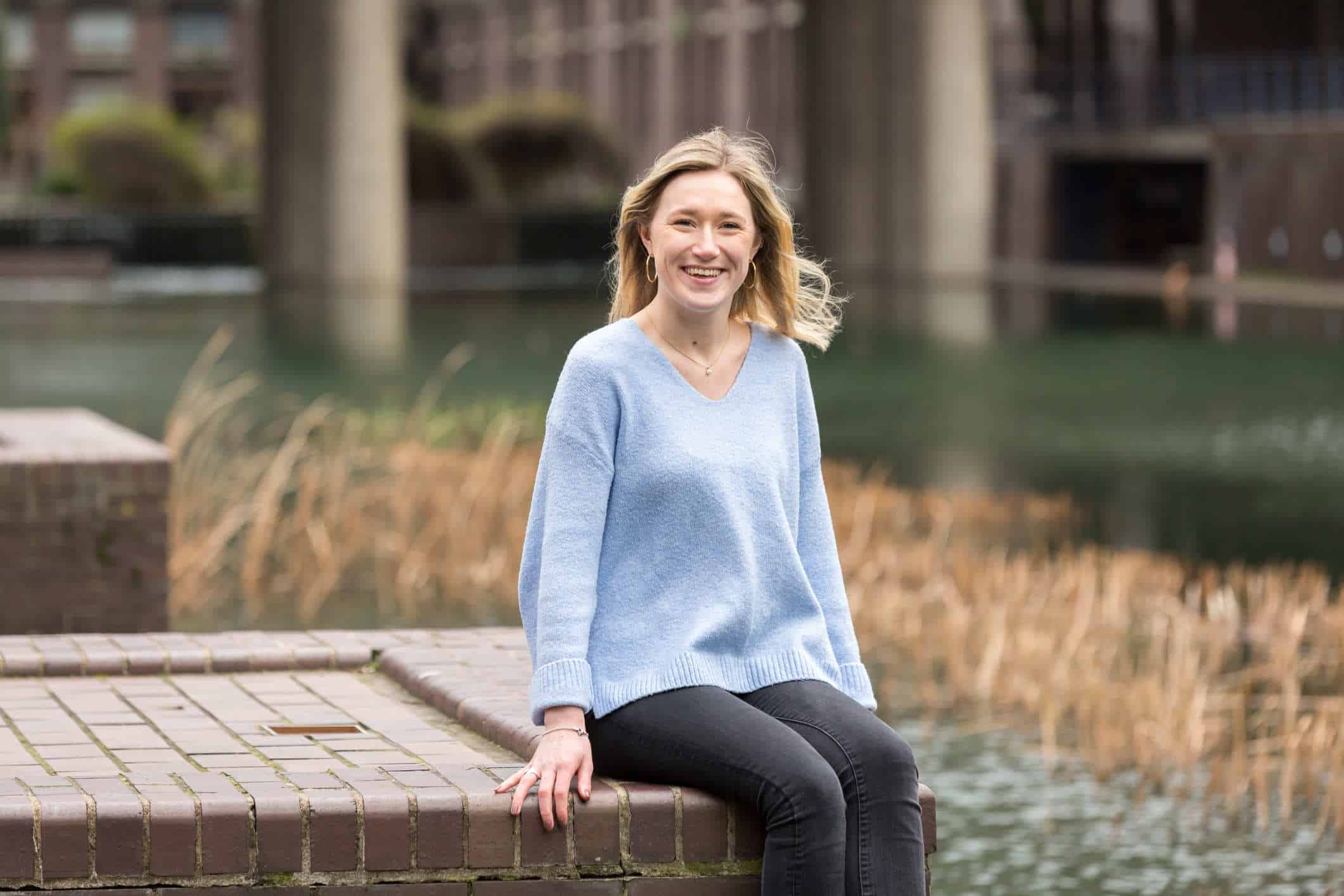 A woman in a blue v-neck jumper is sat on the edge of a wall surrounded by a pond.