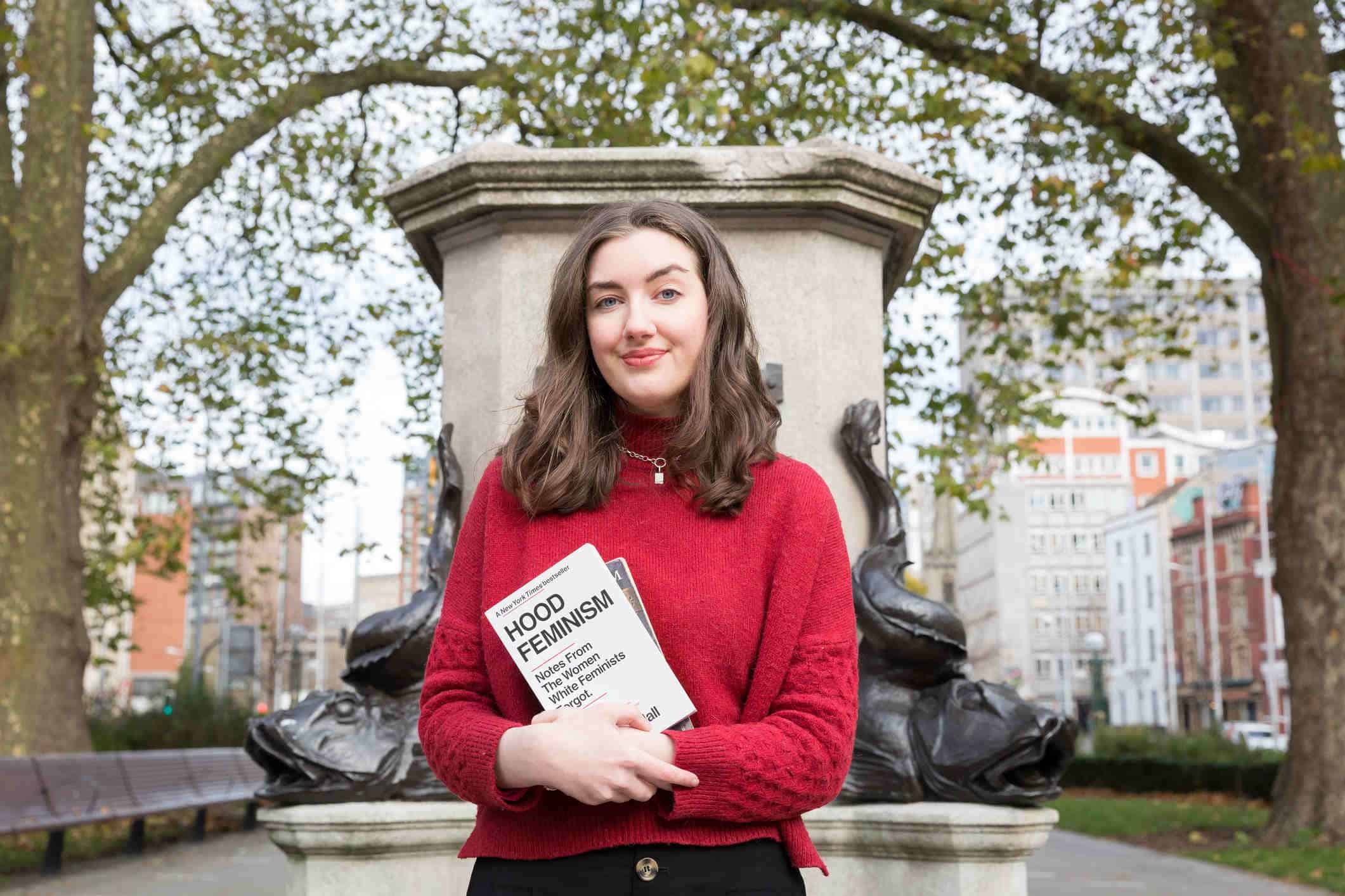 Miranda stands in front of an empty plinth. She is carrying to books, one is titled "Hood Feminism".